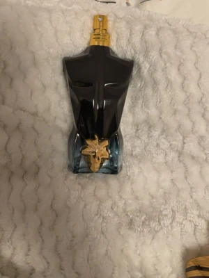 Le Male Le Parfum Jean Paul Gaultier - Unik parfymflaska formad som en manlig torso i svart och blått glas med gulddetaljer och en dekorativ guldblomma framtill. Flaskan har en lyxig och modern design som verkligen sticker ut i badrumshyllan.