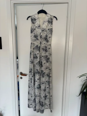 Vit långklänning med svart mönster - Säljer en ärmlös vit långklänning med svart abstrakt mönster. Klänningen har v-ringning och markerad midja med knytband. Perfekt för dig som gillar en stilren men ändå unik look.