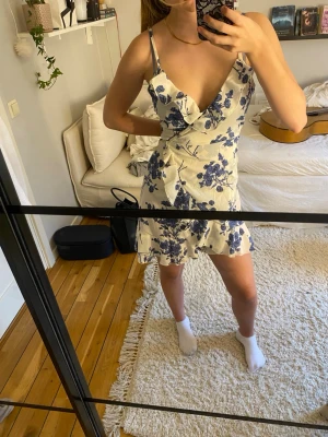 Blommig kortklänning med volang - Söt kortklänning i vitt med blått blommönster och volangdetaljer vid urringningen. Klänningen har smala axelband och en lätt utsvängd kjol som ger en feminin siluett. Perfekt för varma dagar och somriga tillfällen.