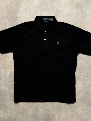 Svart pikétröja från Polo Ralph Lauren - Klassisk svart pikétröja från Polo Ralph Lauren med röd broderad logga på bröstet. Tröjan har korta ärmar, krage och två vita knappar framtill. Tillverkad i bomull för en bekväm och stilren look.