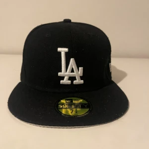Svart LA Dodgers keps New Era 59FIFTY - Klassisk svart LA Dodgers keps från New Era 59FIFTY med vit broderad LA-logga framtill och 1988 World Series-detalj på sidan. Kepsen har rak skärm och broderade loggor även på sidan och bak. Perfekt för dig som gillar sportig streetstyle.