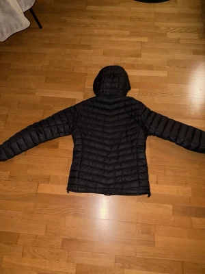 Svart dunjacka med huva från Peak Performance - Säljer en svart dunjacka från Peak Performance med huva och dragkedja framtill. Jackan har en quiltad design och är lätt vadderad för extra värme. Perfekt för kalla vinterdagar och har en stilren look som passar till det mesta.