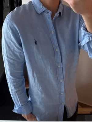 Ljusblå linneskjorta från Ralph Lauren - Säljer en ljusblå skjorta från Ralph Lauren i 100% linne med klassisk krage och knappar framtill. Skjortan har lång ärm och den ikoniska loggan broderad på bröstet. Perfekt för en avslappnad men stilren look. Där är en liten reva på kragen som man ser på sista bilden, men syns knappt när man har den på sig. 