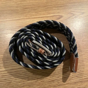 Randigt flätat bälte med läderdetaljer - Snyggt flätat bälte i blått och vitt med randigt mönster. Bältet har detaljer i brunt läder vid spännet och änden, samt ett klassiskt metallspänne. PRUTBART!!!