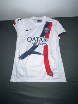 PSG bortatröja Nuno Mendes #25 - Säljer en vit PSG bortatröja från Nike med Nuno Mendes och nummer 25 på ryggen. Tröjan har blå och röda detaljer, Qatar Airways sponsortryck och Champions League-märke på ärmen. Tillverkad i lätt och ventilerande material, perfekt för fotbollsträning eller match.