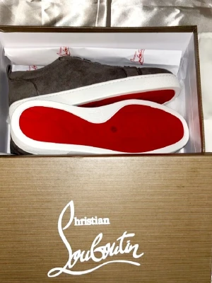 Gråa Christian Louboutin sneakers - Snygga gråa sneakers från Christian Louboutin i mocka med klassisk röd sula och vita kanter. Skorna har rund tå, gråa skosnören och diskret logga på insidan. Perfekta för dig som vill ha en exklusiv och trendig look.