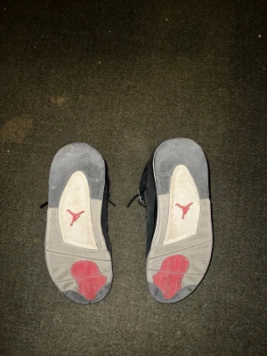 Svarta Air Jordan 4 Retro sneakers - Säljer ett par svarta Air Jordan 4 Retro sneakers med meshpaneler och detaljer i grått och vitt. Sulan har synlig Air-enhet och röda detaljer undertill samt den klassiska Jumpman-loggan. Skorna har en ikonisk siluett och cool design som sticker ut.