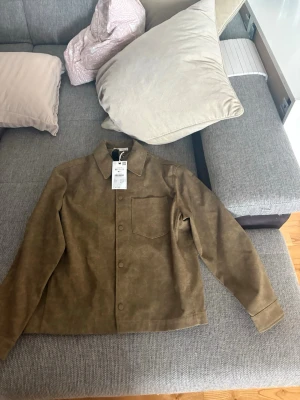 Brun overshirt från Zara - Snygg brun overshirt från Zara i mockaimitation. Klassisk krage, knappstängning framtill och en bröstficka. Perfekt lager på lager-plagg med stilren och enkel design. Passar till många olika outfits och är lätt att matcha.
