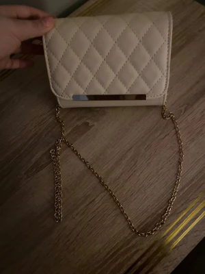 Beige quiltad kuvertväska med kedja - Stilren beige kuvertväska i quiltat konstläder med guldfärgad kedjerem och metall-detalj på framsidan. Väskan har en klassisk rektangulär form och lockstängning. Perfekt för dig som gillar en chic och enkel look.