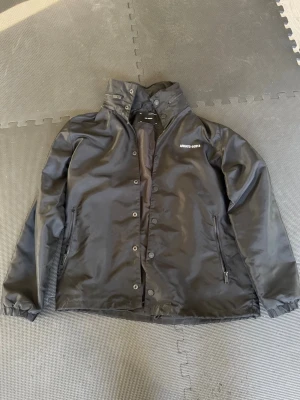 Limiterad! Svart vindjacka från Axel Arigato - Snygg svart vindjacka från Axel Arigato med tryck på bröstet och stor logga på ryggen. Jackan har hög krage, dragkedja och tryckknappar framtill samt två fickor med dragkedja. Materialet är glansigt och vattenavvisande, perfekt för blåsiga dagar. Även en utfällbar luva med hjälp av dragkedja vilket syns på bild. Pris är alltid diskuterbart!