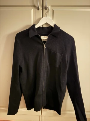 Svart zip-kofta med krage - Stilren svart kofta med dragkedja framtill och klassisk krage. Koftan har en bröstficka på vänster sida och är långärmad. Materialet ser ut att vara mjukt och stickat, perfekt för lager-på-lager. Passar dig som gillar minimalistisk stil.