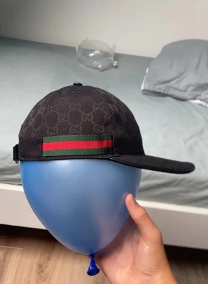 Svart Gucci keps med GG-mönster - Snygg svart keps från Gucci med klassiskt GG-monogram över hela kepsen. På sidan finns en grön och röd rand som ger den en ikonisk look. Justerbar rem bak för perfekt passform. Materialet är troligtvis bomullscanvas.