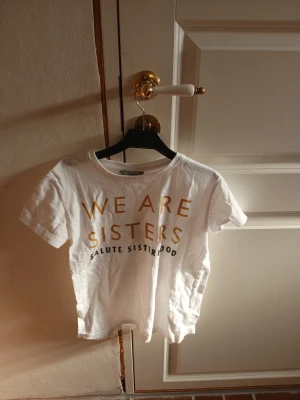 Vit t-shirt med guldtext - Vit t-shirt med korta ärmar och rund hals. Framsidan har trycket 'WE ARE SISTERS SALUTE SISTERHOOD' i guldiga bokstäver. Perfekt för dig som vill ha ett statement-plagg med enkel och clean design.