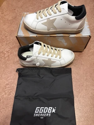 Golden goose sneaker - Snygga vita sneakers från Golden Goose med beige stjärna på sidan, beige skosnören och svart detalj vid hälen. Skorna har en klassisk låg profil och är tillverkade i läder med perforerade detaljer på tungan. Perfekta för en trendig streetwear-look.