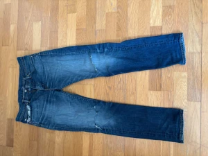 Levi's blå raka jeans - Säljer ett par klassiska blå Levi's jeans med raka ben och slitningar på låret och knät. Jeansen har fem fickor, knappgylf och den ikoniska röda Levi's-lappen på bakfickan. Perfekta för en avslappnad och trendig look.