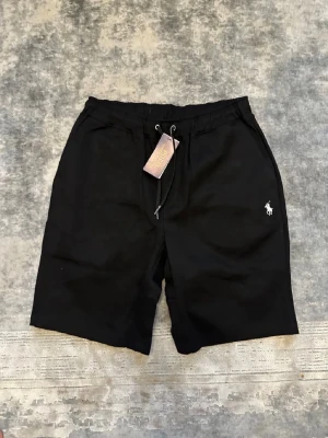 Svarta shorts från Polo Ralph Lauren - Svarta shorts från Polo Ralph Lauren med dragsko i midjan och klassisk logga broderad på vänster ben. Shortsen har sidofickor och en bakficka, perfekt för en avslappnad och stilren look.