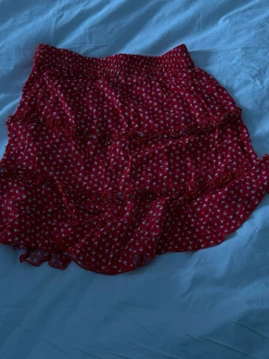 Röd volangkjol från Dobber XS - Supersöt röd kjol från Dobber med små vita blommor och volangdetaljer. Kjolen har resår i midjan och är gjord i ett lätt, luftigt tyg som passar perfekt till sommaren. Den har flera lager volanger som ger en flowy och lekfull look.