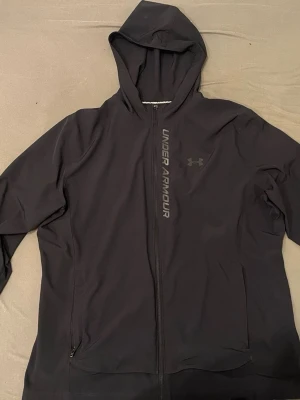 Svart hoodie från Under Armour - Snygg svart hoodie från Under Armour med dragkedja framtill och logga på bröstet. Tillverkad i ett lätt och ventilerande material med meshfoder på insidan. Huva och två sidofickor gör den perfekt för träning eller chill. Stora Under Armour-trycket längs dragkedjan. Dragkedjan saknar själva drag grejen men den går att dra upp ändå 