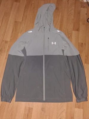 Grå vindjacka från Under Armour - Snygg tvåtonad grå vindjacka från Under Armour med huva och hel dragkedja framtill. Jackan har logga på bröstet, långa ärmar och praktiska fickor. Perfekt för dig som vill ha en sportig och stilren look.