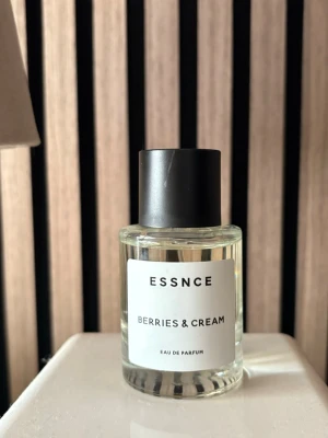 ESSNCE Berries & Cream parfym - ESSNCE Berries & Cream Eau de Parfum i en stilren, genomskinlig glasflaska med svart lock. Doften har en fräsch och söt karaktär med inslag av bär och grädde. Flaskan har en enkel vit etikett med svart text och modern design.