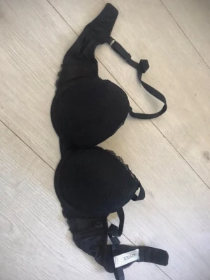 Svart push-up bh från Lindex - Snygg svart push-up bh från Lindex med spetsdetaljer och justerbara axelband. Bh:n har vadderade kupor och en elegant design som ger extra lyft. Märkt med 'Wow' på etiketten och storlek 75A.