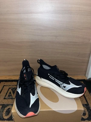 Svarta Mizuno löparskor - Snygga svarta Mizuno sneakers med vit och svart mesh-överdel, vita detaljer och logga på sidan. Skorna har en tjock, vit sula och orange detalj på tån. Perfekta för dig som gillar sportig och modern stil.