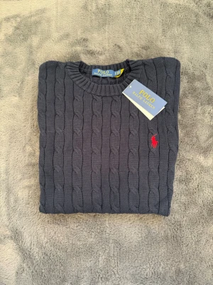 Mörkgrå kabelstickad tröja Polo Ralph Lauren - Snygg mörkgrå kabelstickad tröja från Polo Ralph Lauren med klassisk röd logga broderad på bröstet. Tröjan har rund halsringning och ribbade muddar. Perfekt för dig som gillar stilrena och tidlösa plagg.