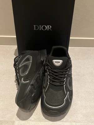 Dior B30 Svarta - Snygga svarta Dior B30 sneakers med reflekterande detaljer och meshpaneler. Skorna har en chunky sula och vita kontrastsömmar som ger en modern vibe. Klassisk snörning och logga på plösen. Perfekta för dig som gillar exklusiva och trendiga sneakers.