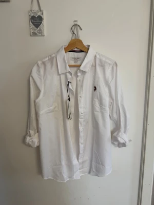 Vit skjorta från U.S. Polo Assn - Klassisk vit skjorta från U.S. Polo Assn med broderad logga på bröstet. Skjortan har lång ärm, knappar framtill och en avslappnad passform. Tillverkad i bomull för en skön känsla och stilren look.