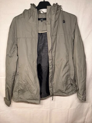 Grå vindjacka med huva och dragkedja - Stilren grå vindjacka med hel dragkedja framtill och justerbar huva med snörning. Jackan har en enkel design och är perfekt för blåsiga dagar. Tillverkad i syntetmaterial och har långa ärmar för extra skydd.