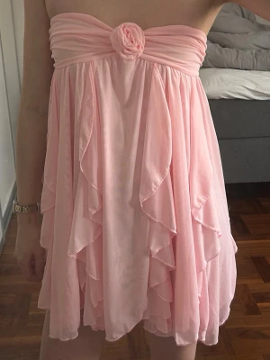 Ljusrosa volangklänning med rosett - Supersöt ljusrosa kortklänning med volanger och en stor rosett framtill som aldrig är använd och har kvar lapparna. Axelbandslös modell med flowy tyg!!