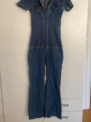 Blå jeansjumpsuit med kort ärm - Snygg blå jeansjumpsuit med korta ärmar,  knappar framtill och lite bootcut style. Aldrig använt!
