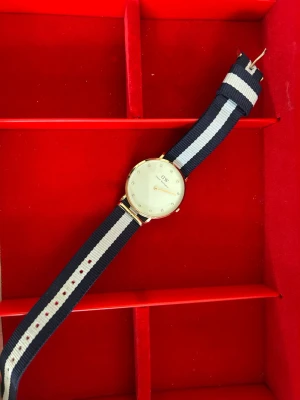 Randig klocka från Daniel Wellington - Snygg klocka från Daniel Wellington med marinblått och vitt randigt tygarmband. Urtavlan är vit med guldfärgad boett och glittriga markeringar istället för siffror. Perfekt accessoar för dig som gillar stilrena detaljer.