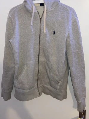 Grå zip hoodie från Polo Ralph Lauren - Klassisk grå hoodie med dragkedja från Polo Ralph Lauren. Tröjan har huva med vita snören, känguruficka framtill och den ikoniska mörkblå loggan broderad på bröstet. Tillverkad i mjuk bomullsblandning för maximal komfort. dragkedjan är av men är fortfarande fullt fungerande storlek S/XS