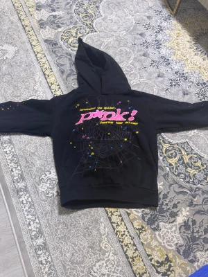 Svart hoodie med tryck och stjärna - Cool svart hoodie från Sp5der! med färgglatt tryck på ryggen och texten 'Pink!' i rosa samt gula detaljer. Stor spindelnätsgrafik och små stjärnor ger en unik look. På framsidan finns en gul stjärna tryckt upptill. Perfekt för dig som gillar streetwear och grafiska motiv.