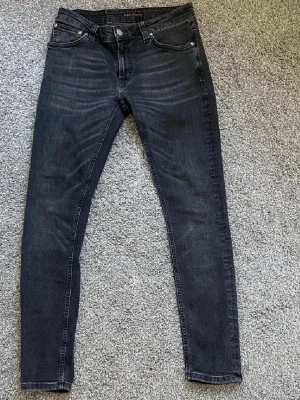 Svarta jeans från Nudie Jeans - Säljer ett par svarta jeans från Nudie Jeans med klassisk femficksdesign och diskreta slitningar framtill. Jeansen har låg midja och smal passform hela vägen ner, tillverkade i stretchig bomull för extra komfort. Perfekt för dig som gillar en tajt siluett.