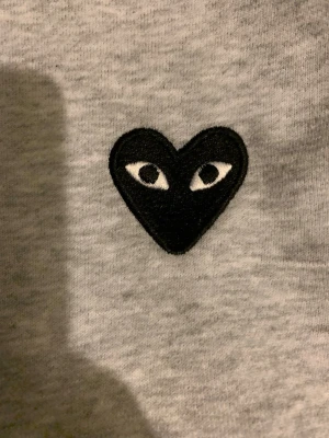 Grå hoodie från Comme des Garçons Play - Snygg grå hoodie med dragkedja från Comme des Garçons Play. Har det ikoniska svarta hjärtat med ögon broderat på bröstet. Utrustad med huva och dragsnören samt två fickor framtill. Tillverkad i mjukt bomullsmaterial för skön känsla.