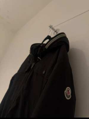 Svart vindjacka från Moncler - Snygg svart vindjacka från Moncler med dragkedja, huva och ribbade muddar med vita ränder. Jackan har två bröstfickor med knappar och Moncler-logga på ärmen. Tillverkad i lätt, vindtätt material som passar perfekt till de flesta stilarna.