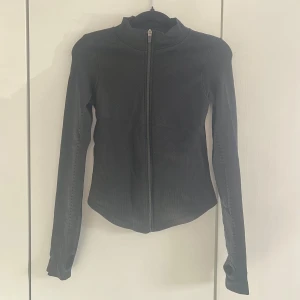Svart ziptröja  - Snygg svart tight ziptröja med hög krage och långa ärmar. Tröjan har figurnära passform och tumhål vid ärmsluten för extra stilpoäng. Perfekt till lager-på-lager och enkel att matcha med det mesta. Materialet är stretchigt och bekvämt.