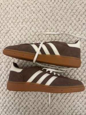 Adidas Spezial bruna sneakers - Bruna Adidas Spezial sneakers i mocka med klassiska vita ränder på sidorna och vit häl. Skorna har en retro look med låg profil och gummisula i brun färg. Snörning framtill och diskret Adidas-logga på sidan. Perfekta för en avslappnad streetwear-stil.