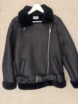Svart pälsjacka från NA-KD - Snygg svart pälsjacka från NA-KD med bred krage i fuskpäls, asymmetrisk dragkedja och bälte i midjan. Jackan har två snedställda fickor och mjukt foder. Perfekt för kalla dagar och ger en cool look.