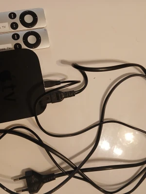 Apple TV med fjärrkontroller och kablar - Apple TV mediaspelare i svart med två silverfärgade Apple TV-fjärrkontroller och tillhörande strömkabel samt HDMI-kabel. Smidig lösning för streaming och underhållning hemma. Klassisk Apple-design i aluminium och plast.