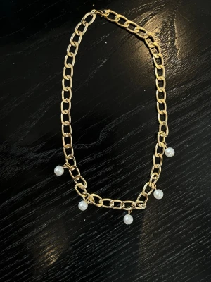 Guldfärgat halsband med pärlor - Snyggt halsband med grov guldfärgad kedja och fem vita pärlor som hänger längs länkarna. Kedjan har en chunky design och pärlorna ger en elegant touch. Perfekt accessoar för att lyfta din outfit.