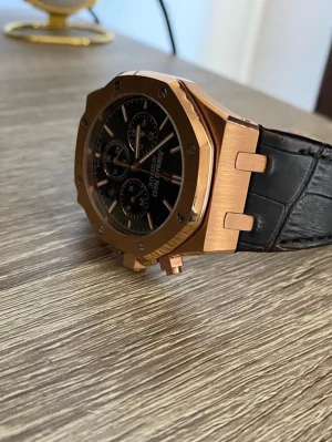 Audemars Piguet Royal Oak klocka guld - Snygg Audemars Piguet Royal Oak klocka med åttakantig boett i guldton och svart urtavla med flera subdials. Svart armband i skinnimitation med viklås i matchande guldton. Perfekt accessoar för dig som gillar exklusiv stil.