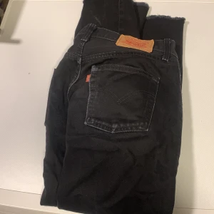 Svarta Levi's jeans med råa benslut - Svarta jeans från Levi's med klassisk femficksdesign och råa, avklippta benslut. Jeansen har en tydlig läderpatch bak i midjan och orange Levi's-tag på bakfickan. Perfekta för en avslappnad och trendig look. Passade ej mig! Storlek W32 L30