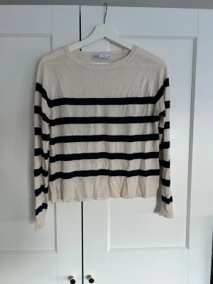 Randig stickad tröja från Zara - Beige tunn stickad tröja från Zara med svarta horisontella ränder. Tröjan har rund halsringning, slits i sidan, långa ärmar och ribbade muddar. Materialet är tunt och mjukt. Sömmen i ena slitsen har gått upp lite (se bild 2)