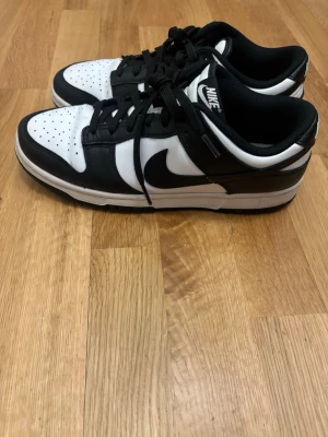 Nike Dunk Low svart/vit - Nike Dunk Low sneakers i svart och vitt läder med klassisk swoosh-logga på sidan. Skorna har perforerad tåbox, svarta skosnören och låg siluett. Perfekta för dig som gillar stilrena och tidlösa sneakers.