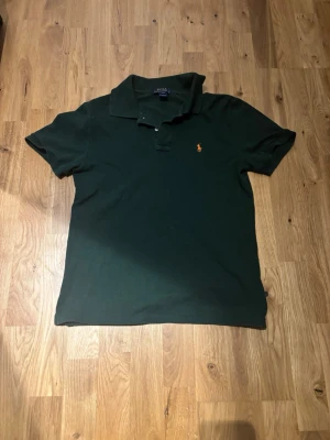 Mörkgrön pikétröja från Ralph Lauren - Klassisk mörkgrön pikétröja från Ralph Lauren med korta ärmar och krage. Tröjan har den ikoniska broderade loggan i orange på bröstet och är tillverkad i mjuk bomull. Perfekt till jeans eller shorts för en clean look. Storlek syns på bilden troligen Large för barn