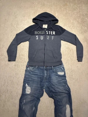 Mörkblå Hollister hoodie med dragkedja ochtryck - Snygg mörkblå hoodie från Hollister med dragkedja och huva. Framtill står det 'Hollister Surf' i vitt tryck och på ärmen finns en brun läderpatch med Hollister-loggan. Hoodien har kängurufickor och är gjord i mjukt bomullsmaterial. Riktigt passlig för dig som vill klä dig chill men ändå se uppklädd ut 