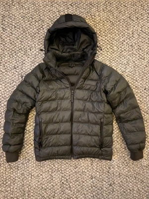 Mörkgrön pufferjacka med huva - Säljer en mörkgrön pufferjacka med huva och dragkedja framtill. Jackan har två sidofickor med dragkedja och ribbade muddar vid ärmsluten. Den quiltade designen ger en klassisk look och materialet är lätt och värmande, perfekt för kalla dagar.